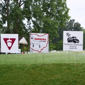 Three signs on a grassy lawn with trees in the background advertise local businesses. The tone is calm and rural, suggesting a community event.