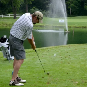 A golfer in a striped shirt and cap swings a club on a lush green course near a pond with a fountain, creating a calm and focused atmosphere.