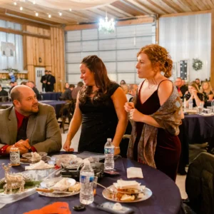 A man seated at a table talks to two women standing beside him in a warmly lit banquet hall. The atmosphere is lively and festive.