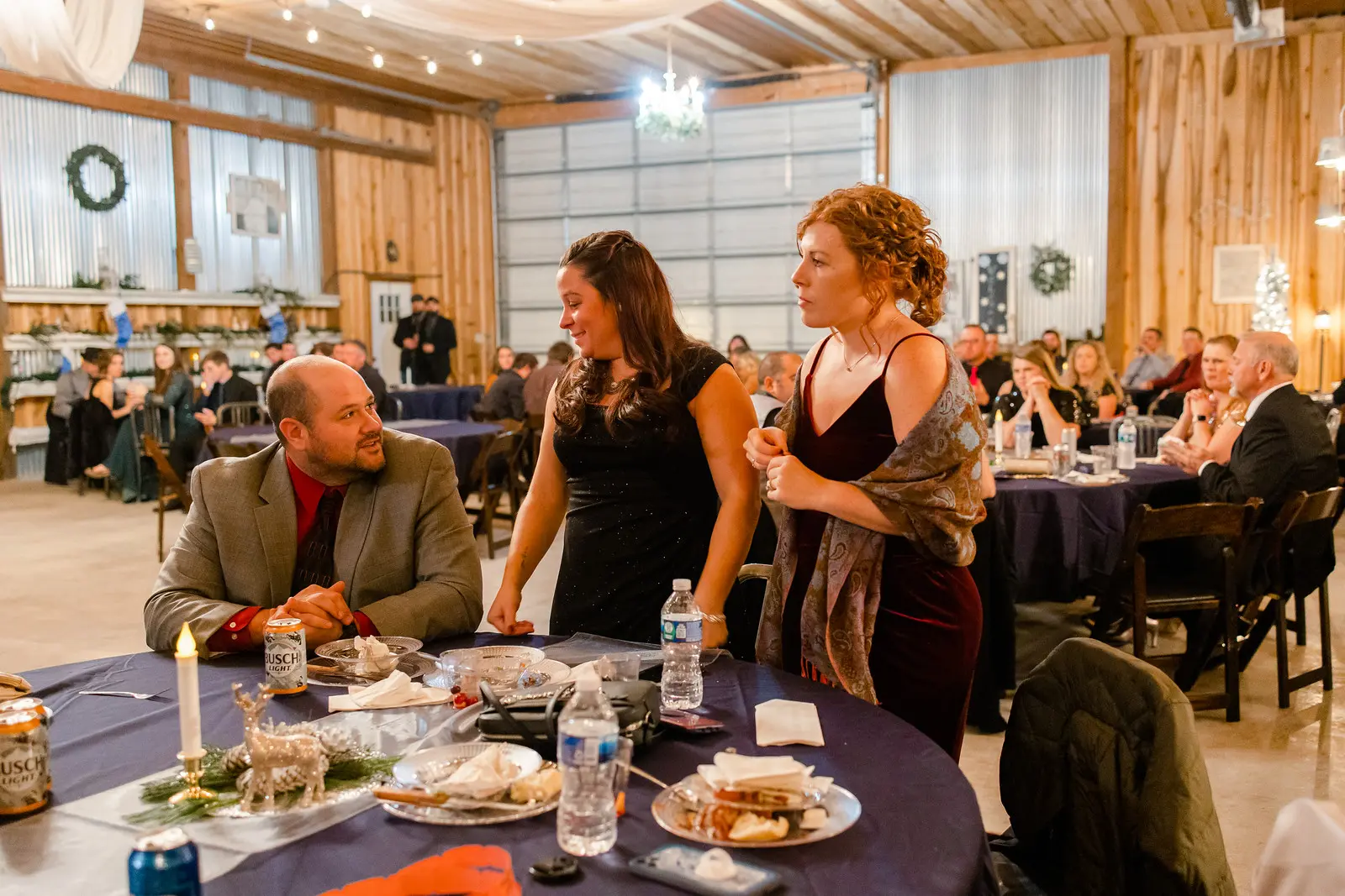 A man seated at a table talks to two women standing beside him in a warmly lit banquet hall. The atmosphere is lively and festive.