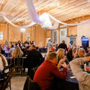 A lively indoor banquet with guests seated at round tables draped in navy cloths. Warm lights and draped fabric create a festive, cozy atmosphere.