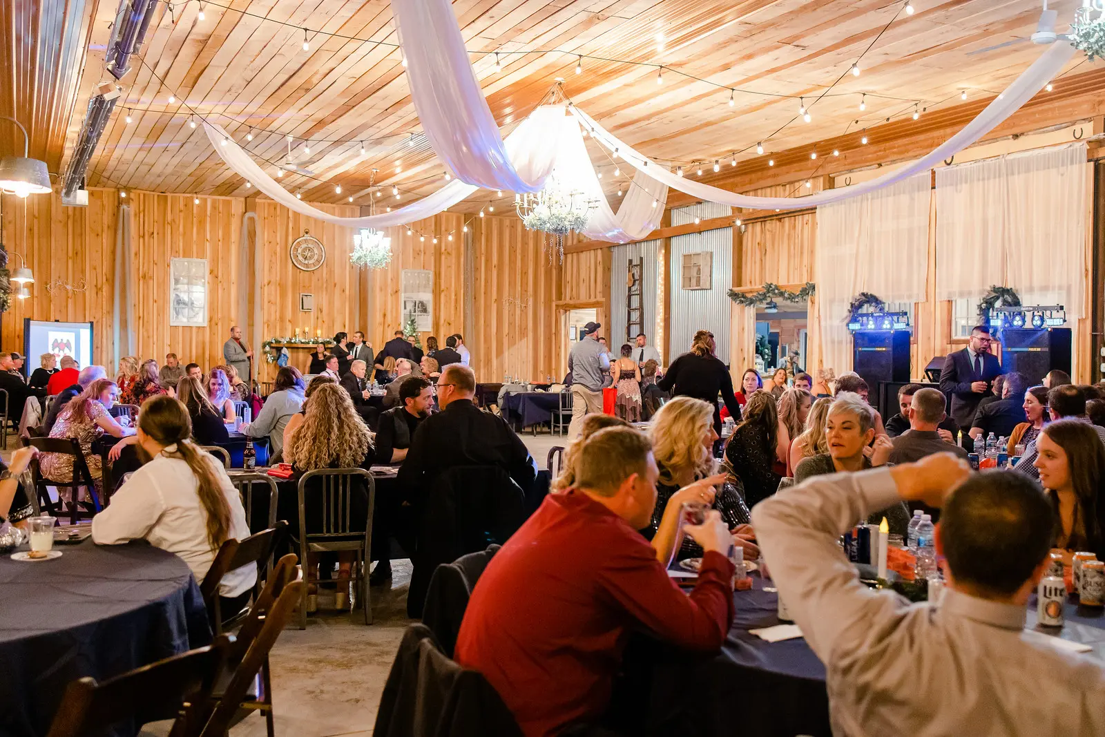 A lively indoor banquet with guests seated at round tables draped in navy cloths. Warm lights and draped fabric create a festive, cozy atmosphere.