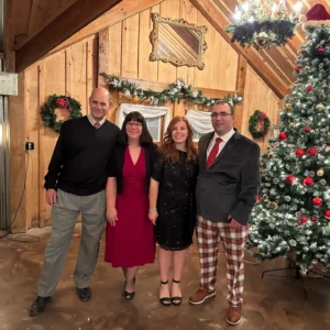 A group of four people smiling indoors, standing by a decorated Christmas tree. The wooden interior creates a cozy holiday atmosphere.