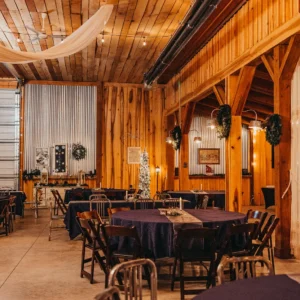 A rustic event space with wooden walls and beams, draped with chiffon. Round tables with navy tablecloths are set, emitting a warm, inviting ambiance.